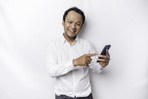 Excited Asian man wearing white shirt smiling while holding his phone, isolated by white background photo
