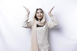 Excited Asian Muslim woman wearing hijab pointing at the copy space above her, isolated by white background photo