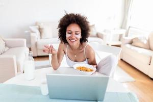 Image of happy young amazing woman sitting indoors at the table with laptop holding corn flakes. Looking at laptop computer and talking to her friends via video call. photo