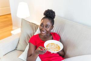 Smiling woman eating healthy breakfast at home in morning, relaxing on couch in living room. Beautiful happy african american girl enjoying food in bowl sitting on sofa photo