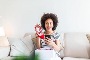 Smiling woman unboxing a postal parcel and taking a selfie with her new purchases using a smartphone. Showing shoes to her friends via video call photo