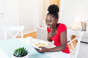 Image of happy young amazing woman sitting indoors at the table with mobile phone holding corn flakes. Looking at screen and talking to her friends via video call. photo