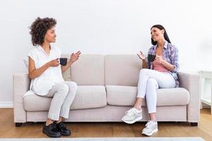 Two female friends in social distancing sitting on sofa in coronavirus pandemic time. Best friends having coffee together while separated by social distancing on sofa at home photo