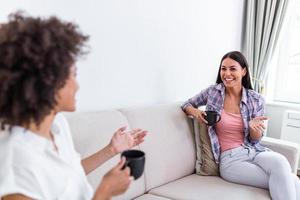 Two female friends in social distancing sitting on sofa. Best friends having coffee together while separated by social distancing on sofa at home photo