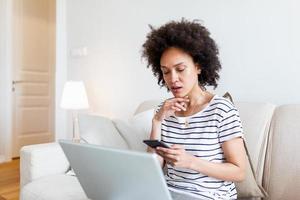 joven negro mujer utilizando teléfono y ordenador portátil a hogar enviar mensajes de texto hermosa mujer estudiando a hogar leyendo un SMS o texto mensaje en su móvil teléfono con un sonrisa como ella mentiras en el sofá con un ordenador portátil foto