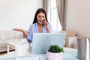 Smiling female multitask working on computer in office talking over phone, happy woman worker using laptop, laughing speaking with friend on cell or communicating with client online, work from home photo