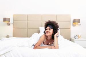 hermosa alegre niña en dormido ojo máscara acostado en cama. joven mujer en cama con dormido máscara sonriente y descansando foto