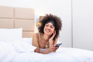 contento niña utilizando un móvil teléfono acostado en el cama a hogar. hermosa joven sonriente mujer acostado en su cama con dormido ojo máscara y utilizando un teléfono en su dormitorio. foto