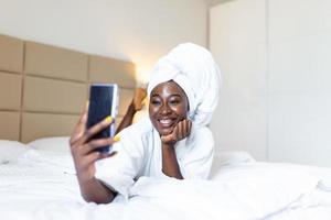 Smiling african young woman lying on bed in bathrobe with her mobile phone taking a selfie. African american woman relaxing on the bed after bath and looking at the phone camera taking a selfie photo