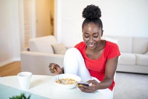 Portrait of young woman is eating cornflakes with milk for breakfast. She is using in mobile phone while sitting at table during eating. photo