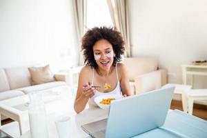 Image of happy young amazing woman sitting indoors at the table with laptop holding corn flakes. Looking at laptop computer and talking to her friends via video call. photo