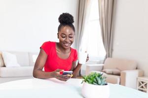 Young woman using a smartphone and having coffee at home. Young african american woman drinking coffee and chatting with friends at social network with her mobile phone photo