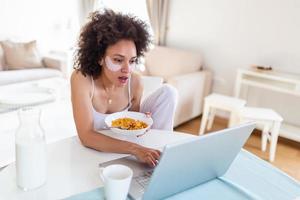 Beautiful young arfican american woman typing on laptop computer at home. Cheerful lady eating corn flakes, drinking morning coffee and working on laptop at home photo