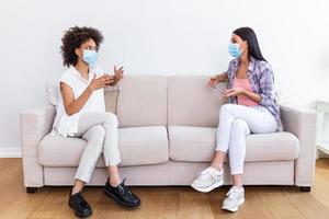 dos hembra mejor amigos sentado en social distancia vistiendo cara máscara y hablando en el sofá, prevenir codicioso 19 coronavirus pandemia infección desparramar. foto
