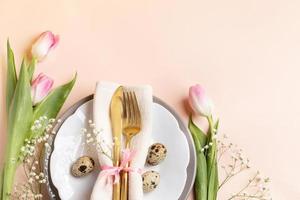 Quail eggs, golden cutlery in napkin on plates, pink tulips, gypsophila on light peach. Copy space. photo