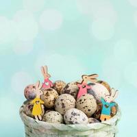 Four small wooden colored Easter bunnies among quail eggs in woven basket on pastel blue. Close up. photo