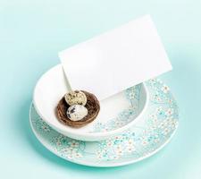 Table setting with Easter quail eggs in nest, white blank sheet on floral pattern plates on blue. photo