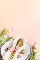 Easter table setting. Quail eggs, golden cutlery, plates, pink tulips, gypsophila on peach backdrop. photo