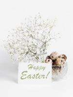 White gypsophila in vase, words Happy Easter on white sheet, quail eggs in crystal bowl on white. photo