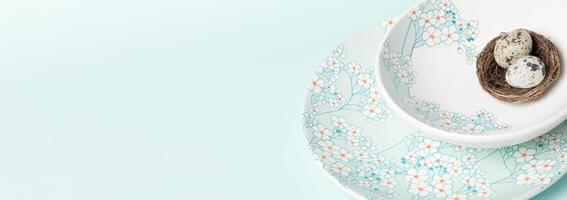 Plates with gentle floral pattern with quail eggs in nest on light blue. Easter banner. Copy space. photo