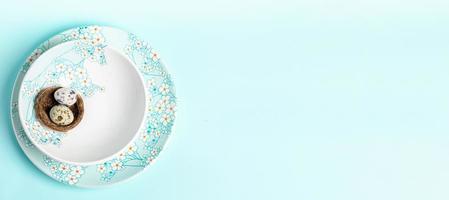 Nest with quail eggs on plates with floral pattern on light blue. Easter banner. Top view Copy space photo