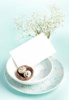 Easter card. Two quail eggs in nest, white empty note on plates with floral pattern on light blue. photo