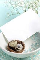 White blank note on plates with floral pattern, quail eggs in nest on light blue Copy space Close up photo