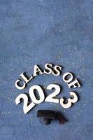 Class of 2023 concept. Wooden number 2023 with graduate hat on dark concrete background with tinsel. photo