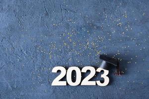 Class of 2023 concept. Wooden number 2023 with graduate hat on dark concrete background with tinsel top view photo