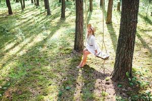 Blond young woman with slender naked legs in white dress and straw hat sitting on a rope swing photo