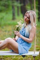 Young beautiful woman in short blue dress posing on a rope swing with bouquet of white flowers photo