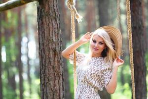 Blond young woman with slender naked legs in short dress and straw hat posing on a rope swing photo