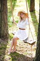 Blond young woman with slender naked legs in white dress and straw hat sitting on a rope swing photo