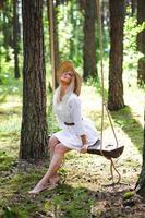 Blond young woman with slender naked legs in white dress and straw hat sitting on a rope swing photo