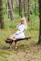 Blond young woman with slender naked legs in white dress and straw hat sitting on a rope swing photo