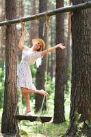 Blond young woman with slender naked legs in short dress and straw hat posing on a rope swing photo