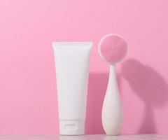 White plastic tube and a massage brush for facial cleansing on a pink background, items for cosmetic procedures photo