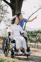 Asian young caregiver nurse support senior older male walking outdoors. Specialist girl doctor help and take care of elderly mature man patient doing physical therapy in public park at nursing home. photo