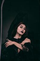 un gótico indonesio mujer vistiendo un hermosa negro sombrero mientras vendaje me gusta un bruja en negro foto