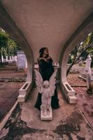 a young witch in a black dress is standing between the tombstones shaped like an angel while visiting the cemetery photo