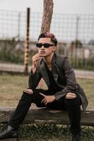 an Asian man with sunglasses is sitting on a tree trunk with a very masculine style in the yard photo