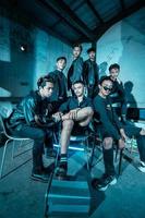 a group of Asian teenagers in black clothes posing very coolly while sitting in a warehouse chair which is lit blue photo