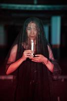 an Asian woman with a transparent veil covered in blood holds a candle in her hand while performing a ritual photo