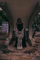 a young witch in a black dress is standing between the tombstones shaped like an angel while visiting the cemetery photo