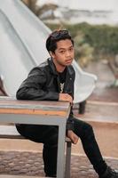 an Asian man is sitting on a wooden chair while wearing a black leather shirt and jacket while stylish with sunglasses photo