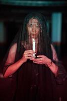 an Asian woman with a transparent veil covered in blood holds a candle in her hand while performing a ritual photo