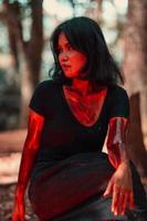 a female serial killer in black clothes and covered in blood has a scary face sitting in the middle of a forest photo