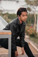 an Asian man is sitting on a wooden chair while wearing a black leather shirt and jacket while stylish with sunglasses photo