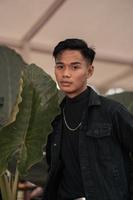 an asian boy in a black denim jacket posing like a naughty boy with a chain around his neck in a park photo