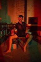 a gay man wearing black and posing with a chair in an old house with a red light photo